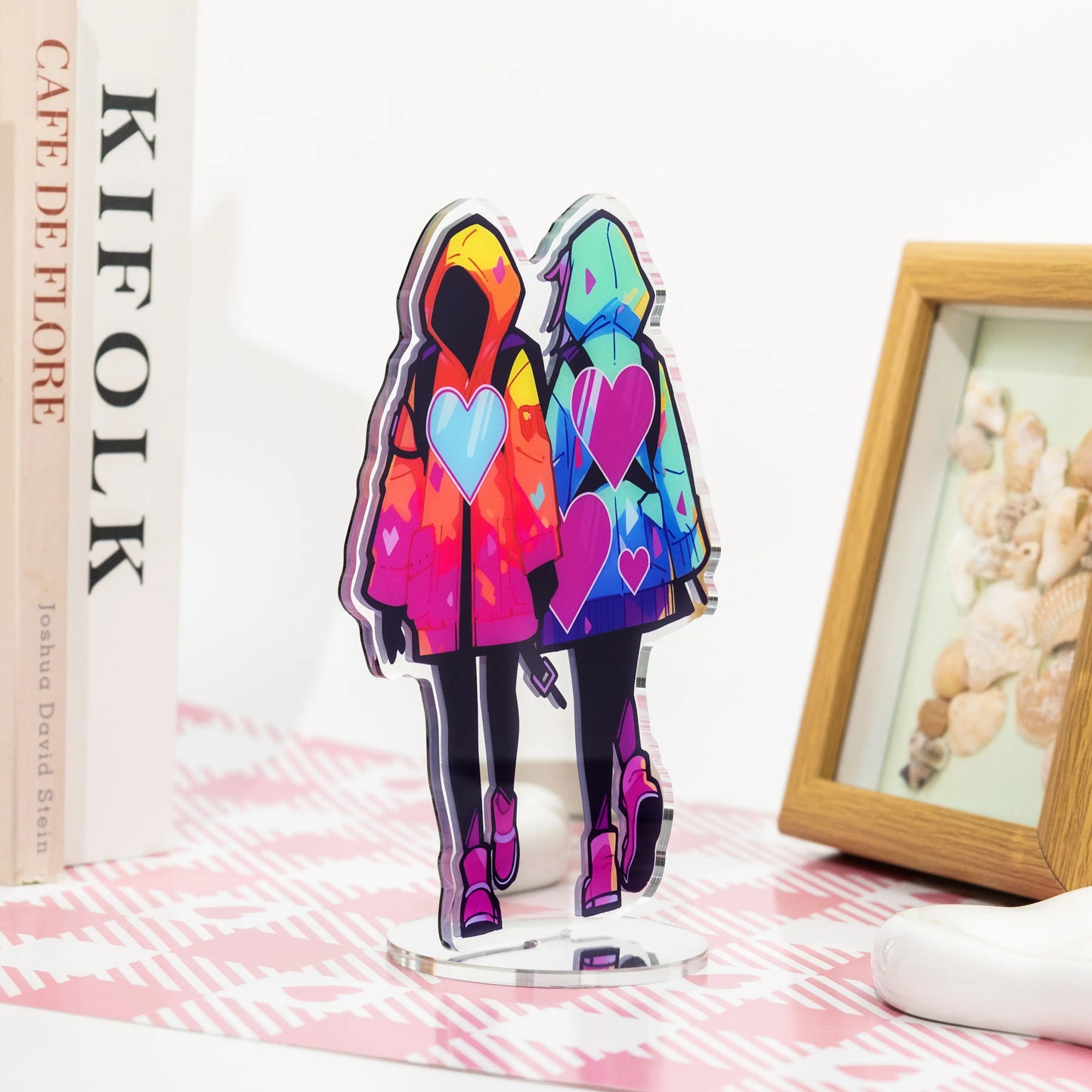 Colorful hooded couple character mirror acrylic standee, anime desk display decor on pink‑white patterned table